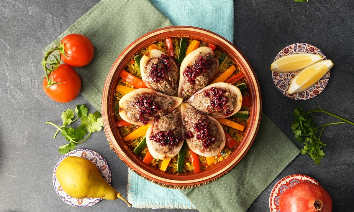 Eine Tajine, gefüllt mit herzhaft gefüllten Birnen. Außen herum liegen Tomaten, Birnen, Zitrone und ein Granatapfel als Deko.