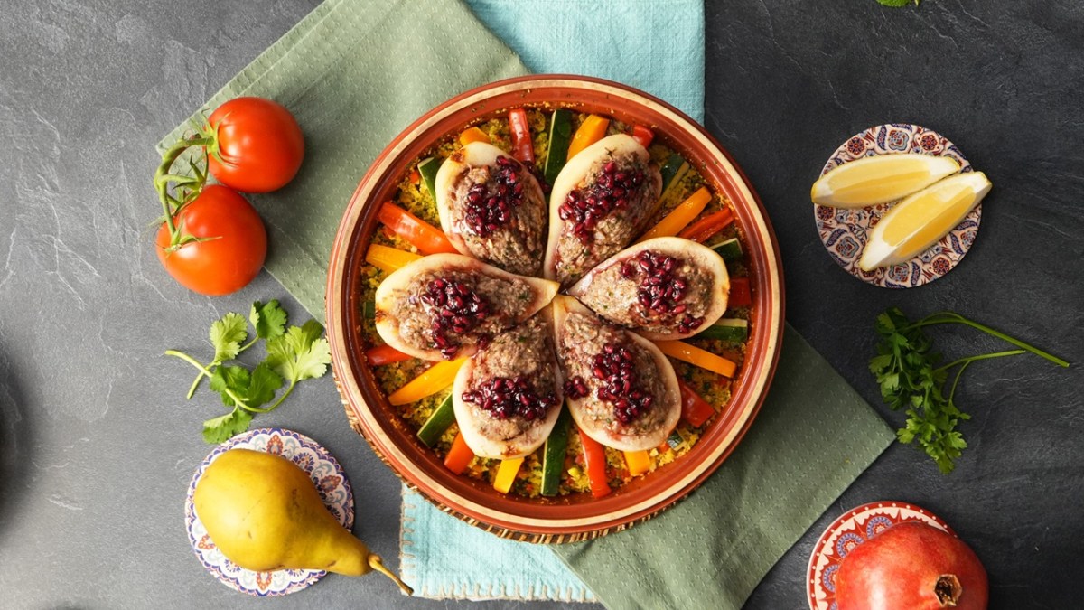 Eine Tajine, gefüllt mit herzhaft gefüllten Birnen. Außen herum liegen Tomaten, Birnen, Zitrone und ein Granatapfel als Deko.