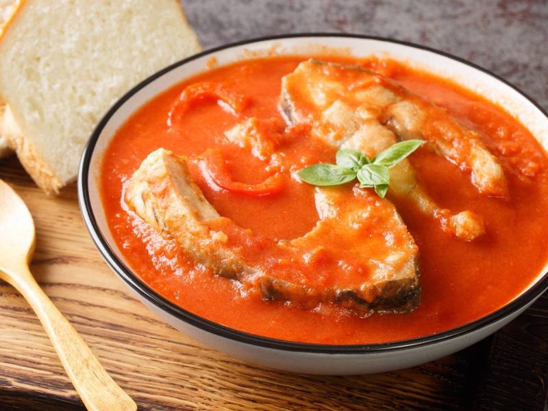 Ein Teller mit ungarischer Fischsuppe. Der Teller steht auf einem Holzbrett und ein Holzlöffel liegt dabei. Die Suppe wird mit zwei Scheiben Weißbrot serviert.