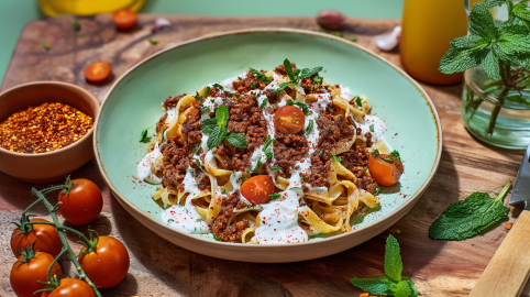 Ein Teller vegetarische Manti Pasta mit Joghurtsoße und frischer Minze. Herum liegen frische Tomaten, frische Minze und Gewürze.