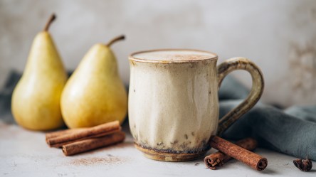 Eine Tasse mit einem warmen Birnen-Chai-Smoothie. Ringsum liegen Zimtstangen und frische Birnen.