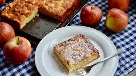 Ein Apfel-Marzipan-Kuchen vom Blech, von dem ein Stück abgeschnitten wurde, welches jetzt auf einem Teller davor liegt.