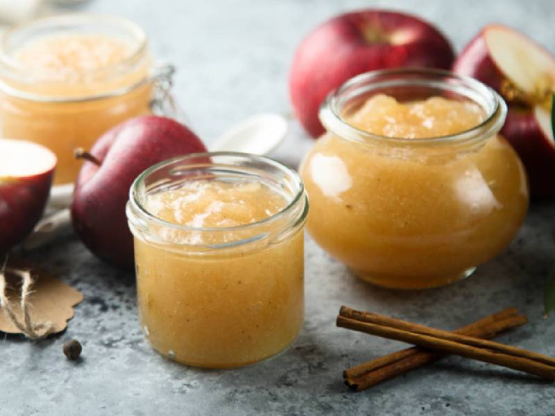 Drei Gläser Apfel-Marzipan-Marmelade auf einem Tisch, Zimtstangen und Äpfel daneben