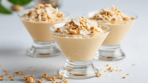 Drei Dessertgläser mit Erdnussbutter-Mousse auf einem weißen Tisch. Sie sind mit Erdnussbutter und weißer Schokolade garniert.