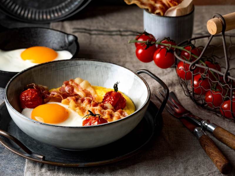 Eine Schale Frühstücks-Polenta mit Ei und Bacon, daneben Kirschtomaten.