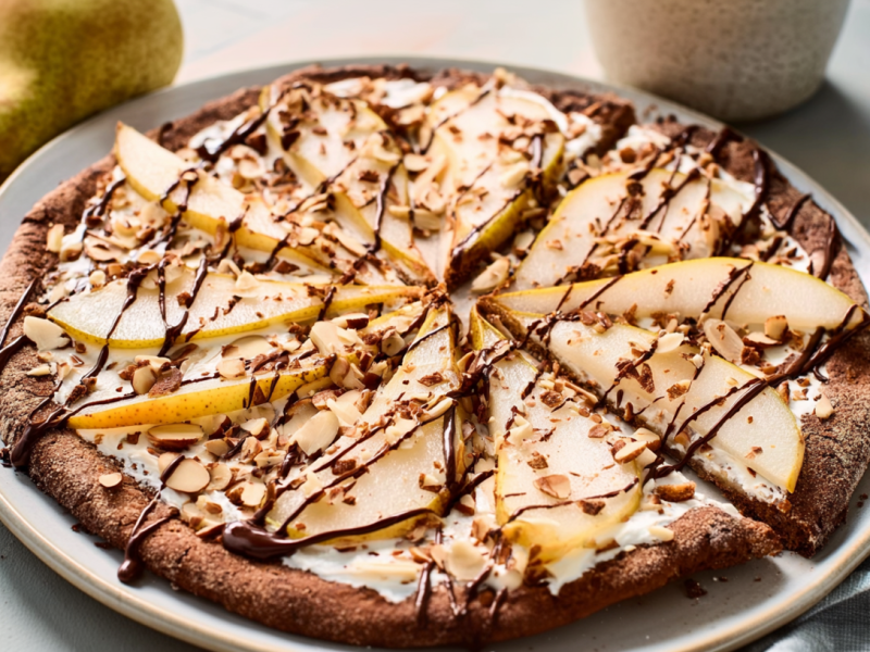 Ein großer Teller mit Frühstückspizza mit Birnen und Mandeln. Im Hintergrund liegen zwei Birnen als Dekoration.