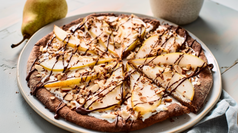 Ein großer Teller mit Frühstückspizza mit Birnen und Mandeln. Im Hintergrund liegen zwei Birnen als Dekoration.