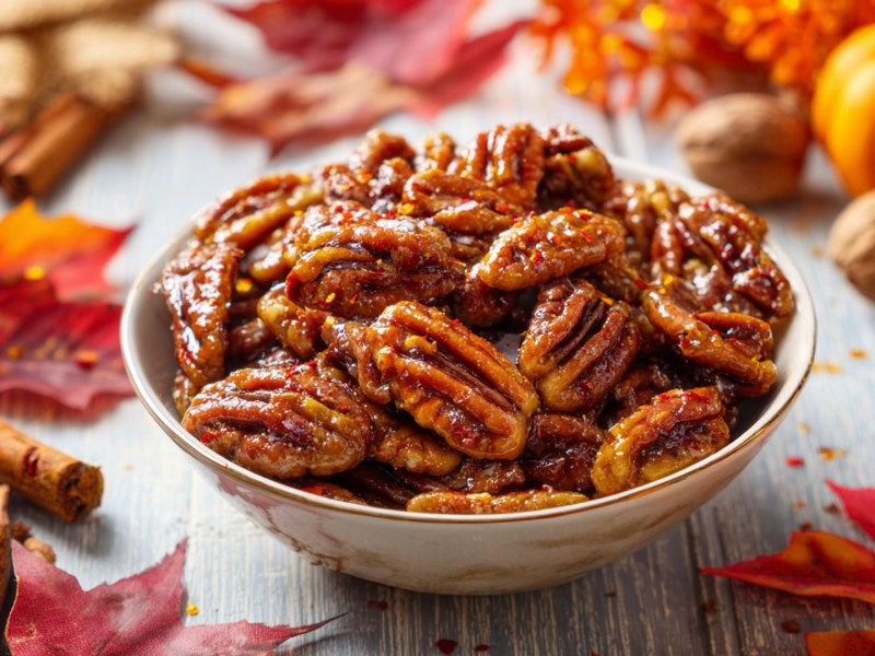 Eine Schüssel voller gerösteter Pekannüsse mit Ahornsirup und Chili. Ringsum liegen Herbstblätter, Zimtstangen und Walnüsse herum.