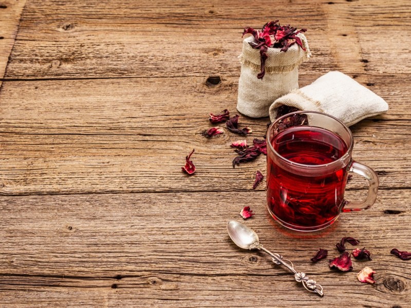 Ein Glas, gefüllt mit Gewürztee mit Hibiskus. Außen herum liegen ein Löffel und Teeblätter als Deko.