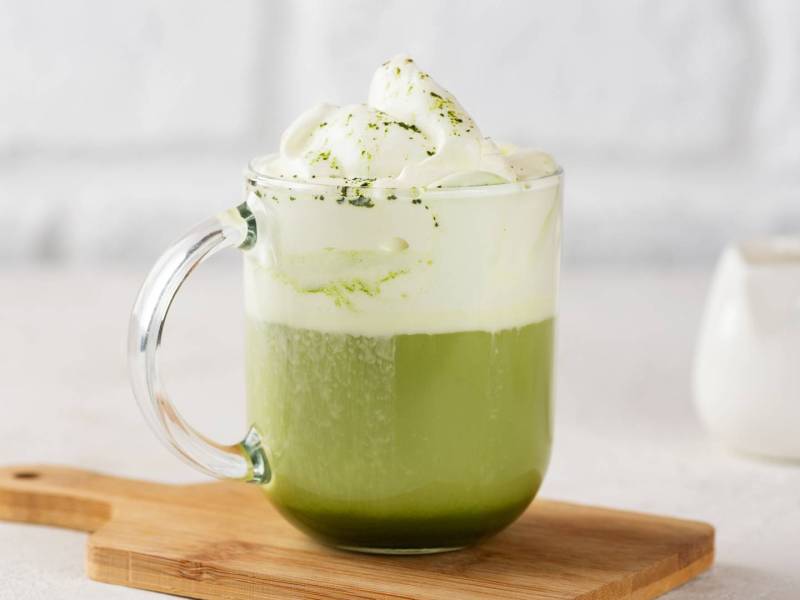 Eine gläserne Tasse heiße weiße Schokolade mit Matcha, die auf einem kleinen Holzbrett steht vor einem hellen, minimalistischen Hintergrund.