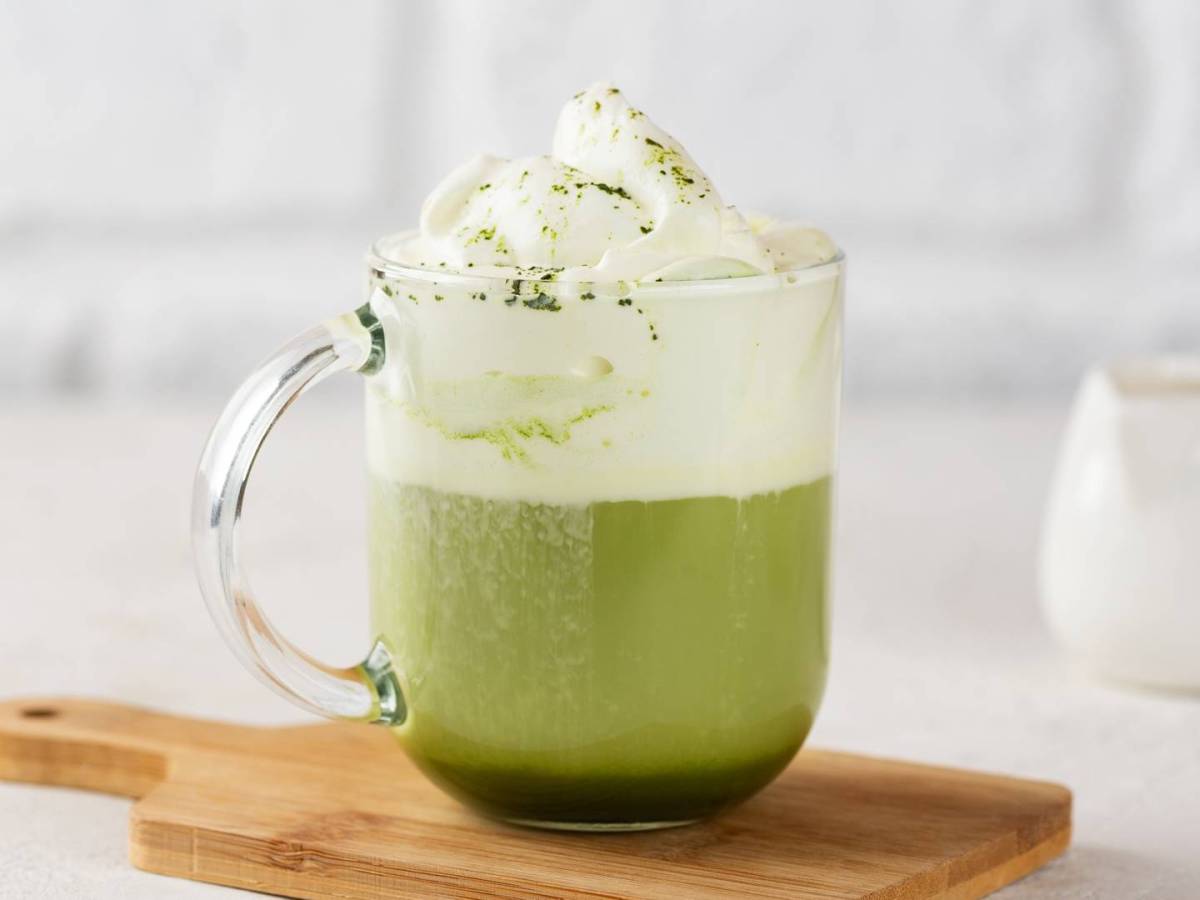 Eine gläserne Tasse heiße weiße Schokolade mit Matcha, die auf einem kleinen Holzbrett steht vor einem hellen, minimalistischen Hintergrund.