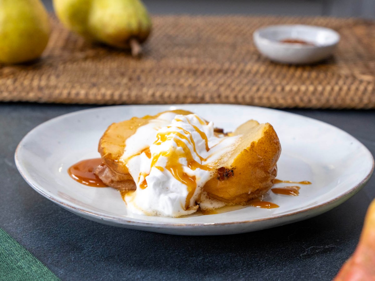Ein Teller mit einer angerichteten holländischen Birne. Im Hintergrund kann man auf einem Tischuntersetzer Birnen und eine Schale mit Gewürz erkennen. Im Vordergrund ragen auch Birnen ins Bild.
