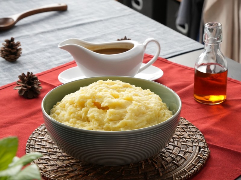 Eine Schüssel mit Kartoffelpüree mit Bratensoße steht auf einem Brett. Ein Tisch ist hübsch gedeckt mit roter Tischdecke und Besteck. Im Hintergrund steht eine Soßenschale.