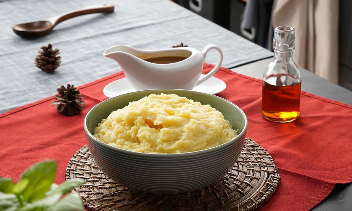 Eine Schüssel mit Kartoffelpüree mit Bratensoße steht auf einem Brett. Ein Tisch ist hübsch gedeckt mit roter Tischdecke und Besteck. Im Hintergrund steht eine Soßenschale.