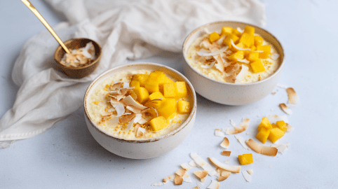 Zwei Schüssel Kokos-Mano-Porridge auf einer grauen Unterlage. Vor ihnen liegen mehrere Kokosraspeln sowie einige Mangowürfel.