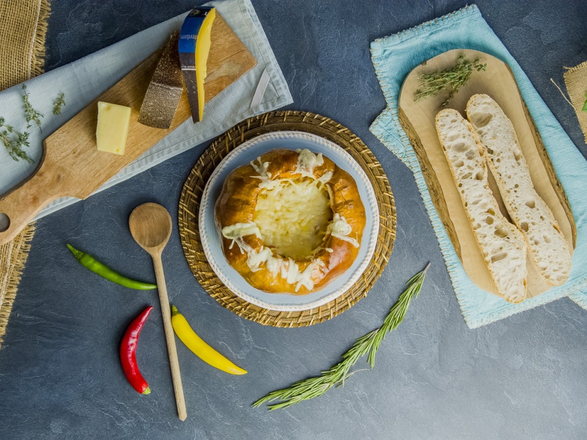 Draufsicht: Kürbis-Käse-Fondue. Daneben liegt ein Brett mit einem aufgeschnittenen Baguette, ein Brett mit Käsestücken sowie ein Holzlöffel und einige Chilischoten verteil.