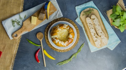Draufsicht: Kürbis-Käse-Fondue. Daneben liegt ein Brett mit einem aufgeschnittenen Baguette, ein Brett mit Käsestücken sowie ein Holzlöffel und einige Chilischoten verteil.