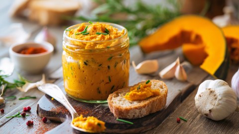 Ein Glas mit Kürbis-Senf-Aufstrich, das umgeben ist von frischen Zutaten wie Kürbisscheiben, einer Knoblauchknolle und Knoblauchzehen, Pfefferkörnern, frischen Kräutern und Brot. Neben dem Glas liegt ein Löffel mit etwas Aufstrich, genauso wie eine Brotscheibe mit etwas Kürbis-Senf-Aufstrich.