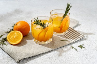 Zwei Gläser mit Mandarinen-Lillet mit Zimt stehen auf einer Stoffserviette. Garniert sind sie mit zwei Rosmarinzweigen. Eine aufgeschnittene Orange und ganze Orange liegen daneben.