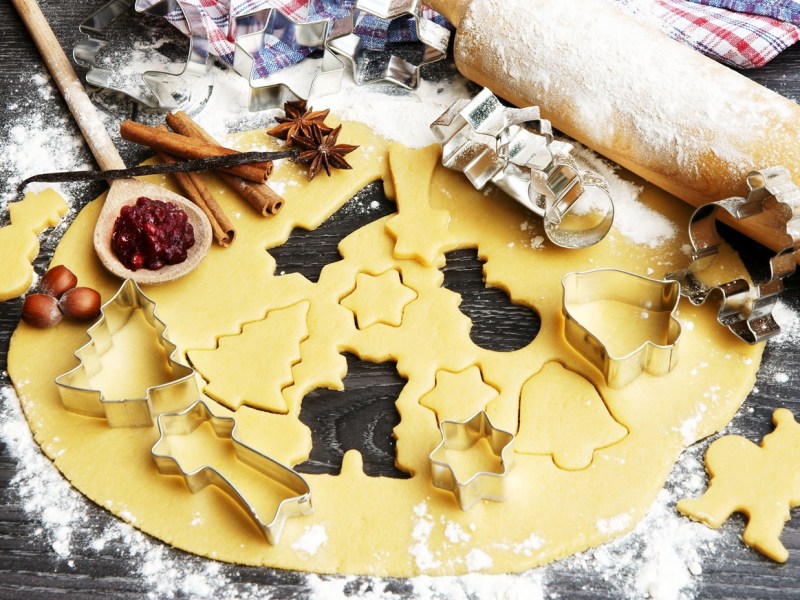 Auf einer bemehlten Arbeitsfläche liegt ein Teig, aus dem einige Plätzchen ausgestochen sind. Daneben liegt ein bemehltes Nudelholz und weiteres Zubehör zum Plätzchenbacken.