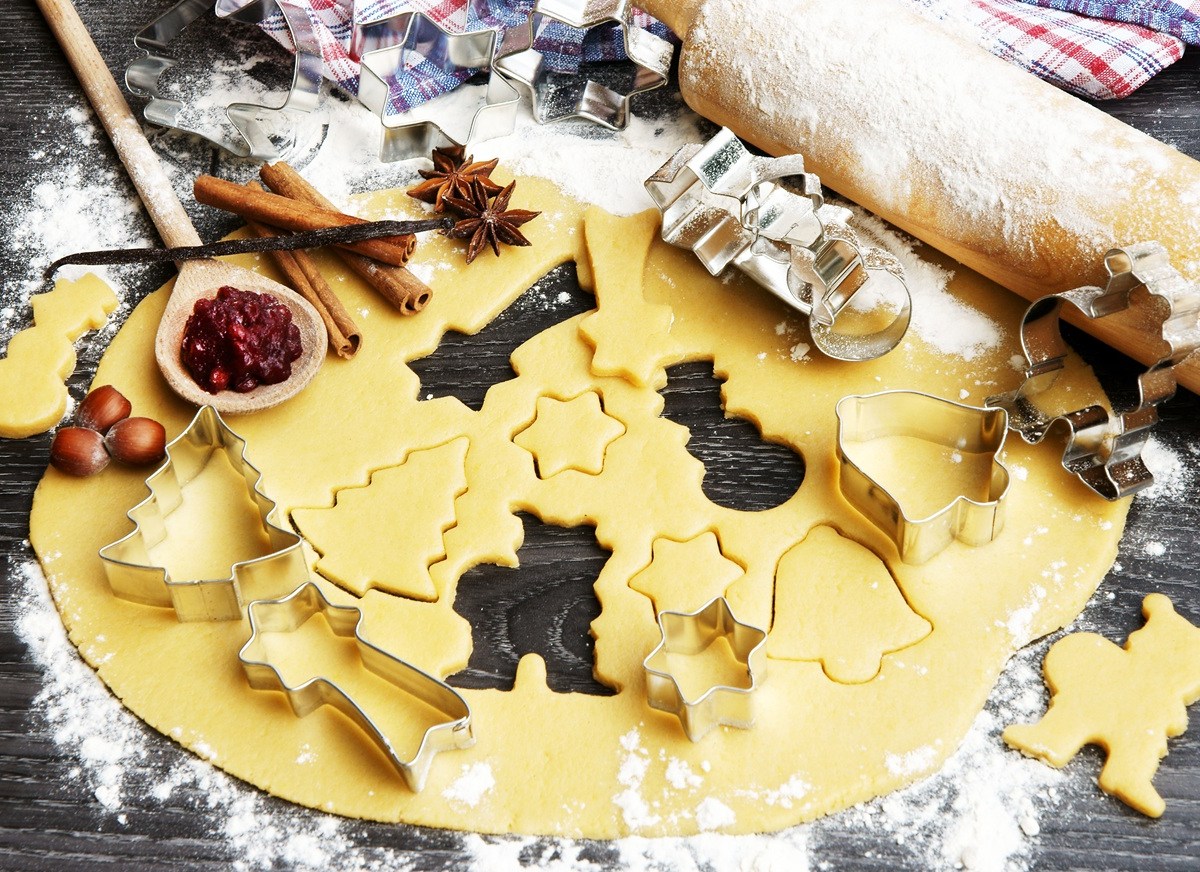 Auf einer bemehlten Arbeitsfläche liegt ein Teig, aus dem einige Plätzchen ausgestochen sind. Daneben liegt ein bemehltes Nudelholz und weiteres Zubehör zum Plätzchenbacken.