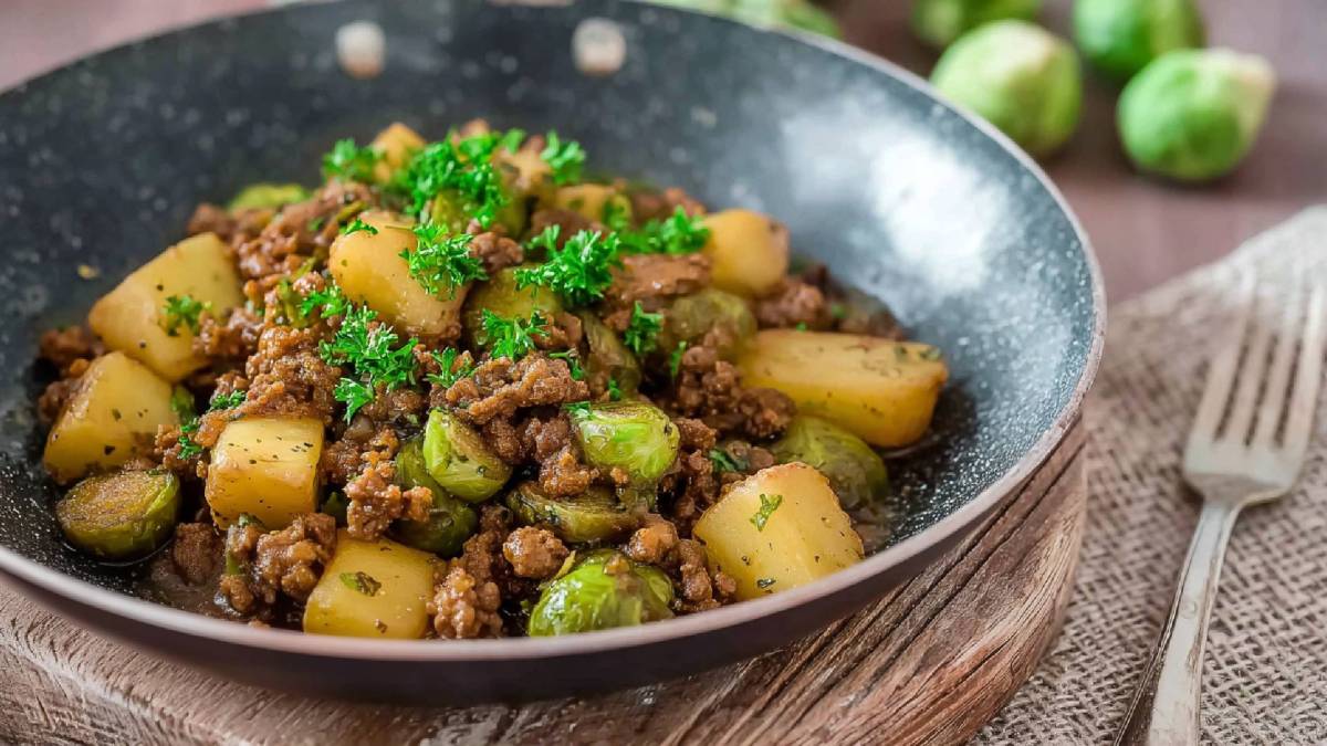 Rosenkohl-Hackfleischpfanne mit Kartoffeln auf einem Tisch, Rosenkohl im Hintergrund und Gabel daneben