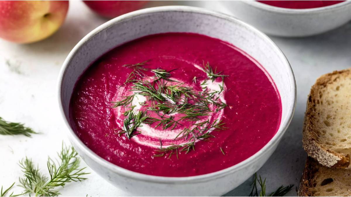 Eine Schale mit Rote-Bete-Cremesuppe mit Apfel auf einem Tisch, Scheibe Brot daneben und Apfel im Hintergrund