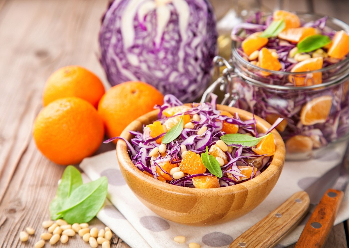 Eine Schüssel voll Rotkohlsalat mit Mandarine. Die Schüssel steht auf einer Stoffserviette. Außen herum liegen Mandarinen und ein Rotkohl. Im Hintergrund steht ein geöffnetetes Bügelglas mit dem Salat.